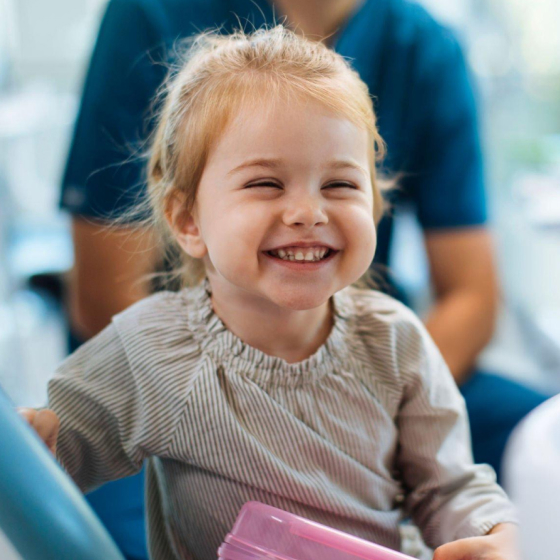 Smiling young girl holding pink folder at pediatric dental office with caregiver in background.