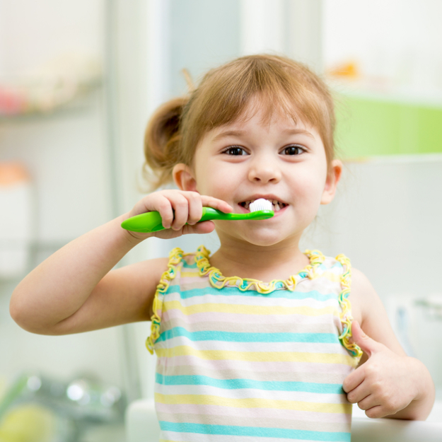 Toddler brushing teeth with green toothbrush in bright, cheerful indoor setting.