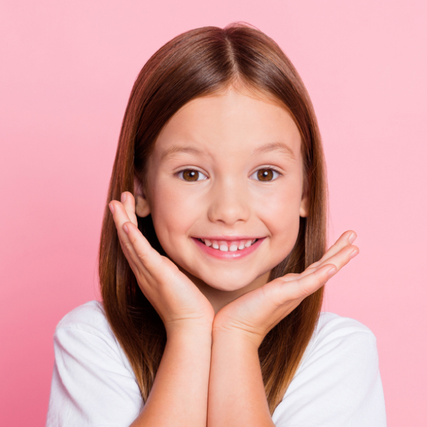 Smiling young girl with hands on face against pink background, pediatric dental care portrait.